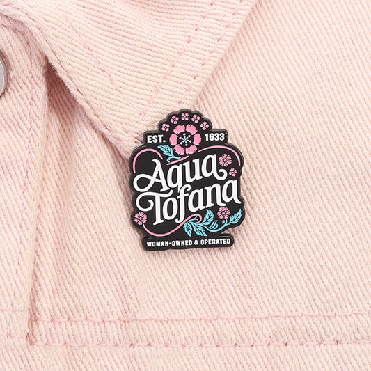 A close-up of a pink woven fabric, likely a denim jacket, featuring a decorative enamel pin that reads 'Aqua Tofana' with pink and teal floral details.