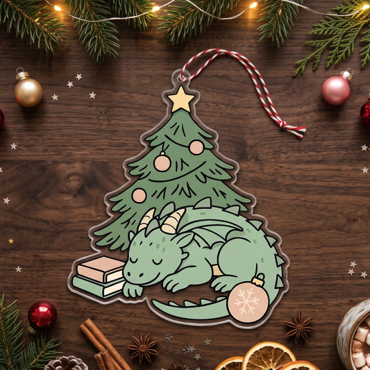 A Christmas-themed die-cut sticker or ornament featuring a sleeping green dragon curled up inside a decorated Christmas tree, surrounded by pine branches, ornaments, ribbons, and holiday decor on a dark wood background.