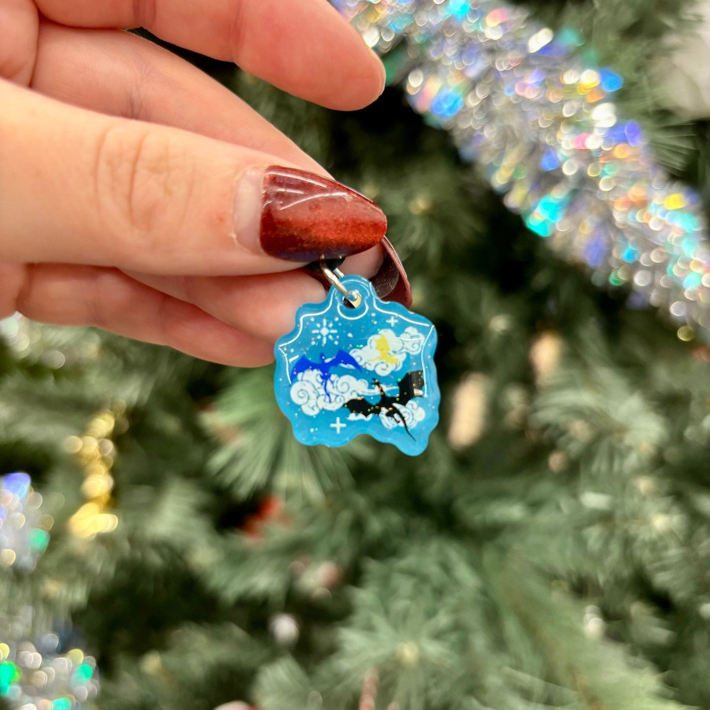 Hand holding a blue kindle charm featuring 3 dragons, one blue, one gold, and one black with a star design against a festive background.