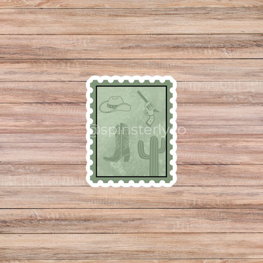 A rectangular postage-stamp-style sticker with a white scalloped border, shown centered on a light brown wood plank background. The sticker has a muted sage green face with western-themed line drawings including a cowboy hat, boots, a revolver, and a cactus.