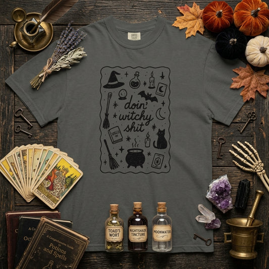 A dark gray graphic t-shirt featuring a black witch-themed illustration with the text 'doin' witchy shit'. The shirt is displayed in a flat-lay arrangement on a dark wooden background, surrounded by autumn-themed props including pumpkins, tarot cards, crystals, old books, and potion bottles.