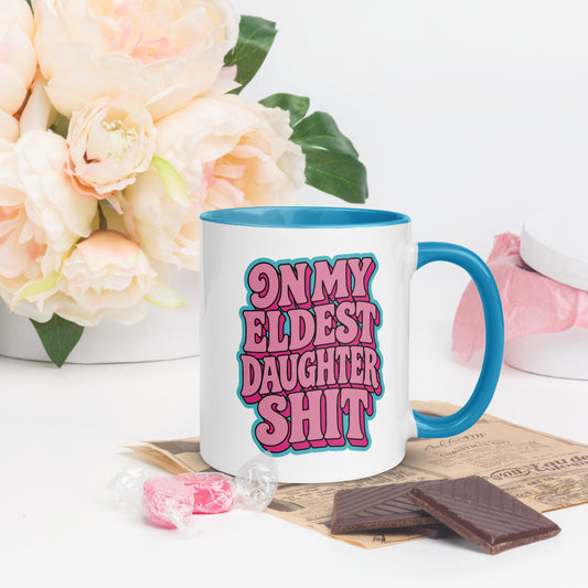 A white ceramic mug with a bright blue handle and interior, featuring the text 'ON MY ELDEST DAUGHTER SHIT' in pink bubble letters with a blue outline. The mug is styled on a white surface with decorative props, including a white vase of pale pink roses, chocolate pieces, and pink candy.