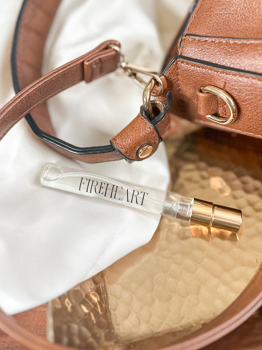 Perfume bottle labeled 'Fireheart' on a reflective surface with a brown leather bag in the background.