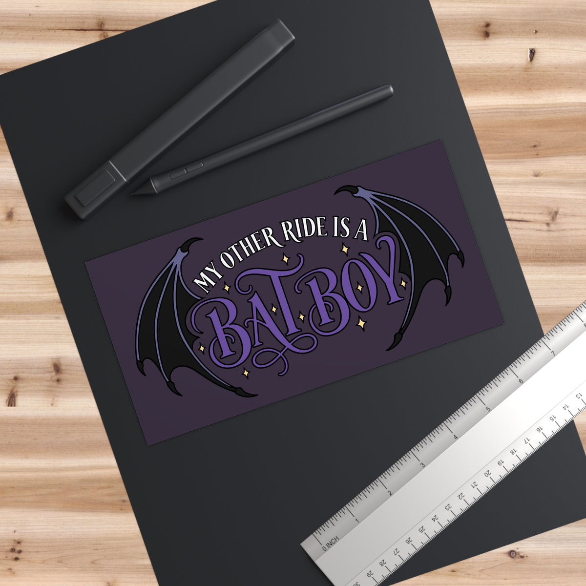 A rectangular printed sticker decal with the phrase “MY OTHER RIDE IS A BATBOY” in stylized purple and white lettering, flanked by black bat wings and small gold stars, shown on a dark sheet over a wood surface with a ruler and stylus nearby.