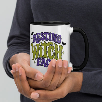 A person holding a white ceramic mug with a black handle and interior. The mug features the text 'RESTING WITCH FACE' in purple and green retro-style lettering with small bat illustrations.