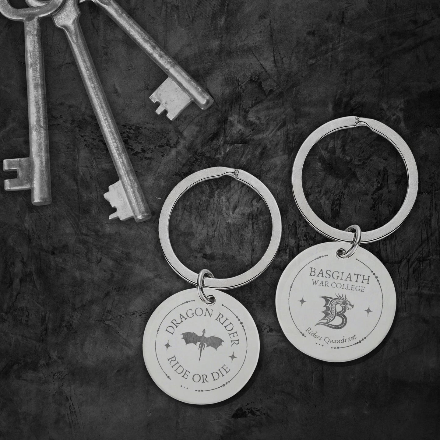 Two engraved silver keychains with round discs hang on a dark textured surface, alongside several antique-style keys. One disc reads “Dragon Rider Ride or Die” with a dragon silhouette, and the other reads “Basgiath War College” with a dragon graphic.