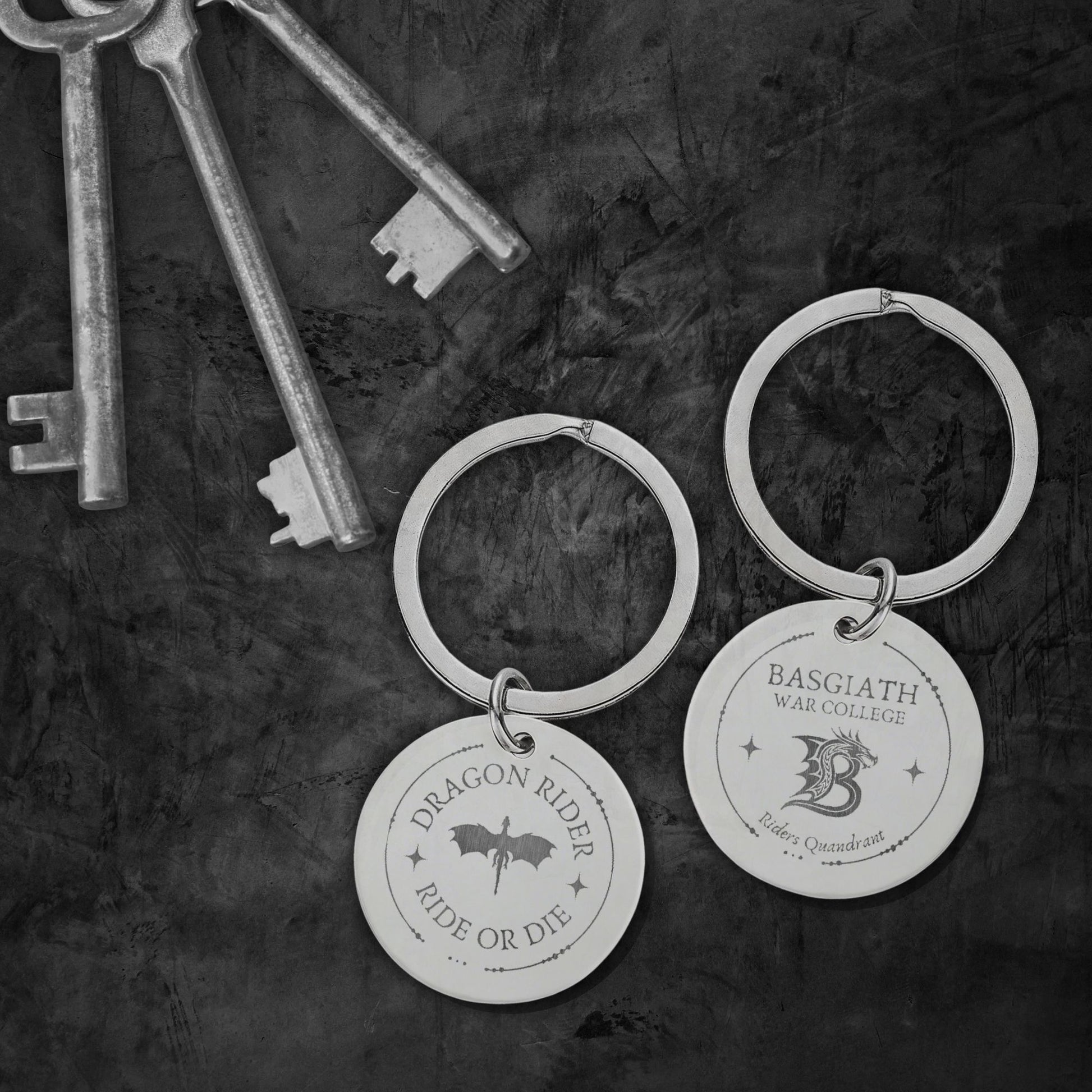 Two engraved silver keychains with round discs hang on a dark textured surface, alongside several antique-style keys. One disc reads “Dragon Rider Ride or Die” with a dragon silhouette, and the other reads “Basgiath War College” with a dragon graphic.