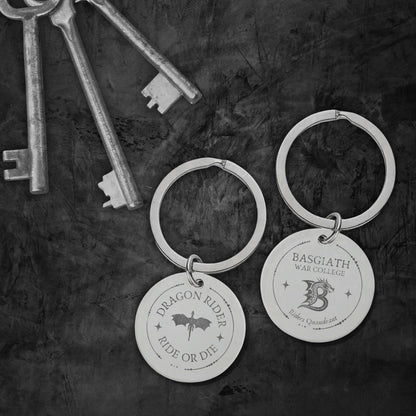 Two engraved silver keychains with round discs hang on a dark textured surface, alongside several antique-style keys. One disc reads “Dragon Rider Ride or Die” with a dragon silhouette, and the other reads “Basgiath War College” with a dragon graphic.