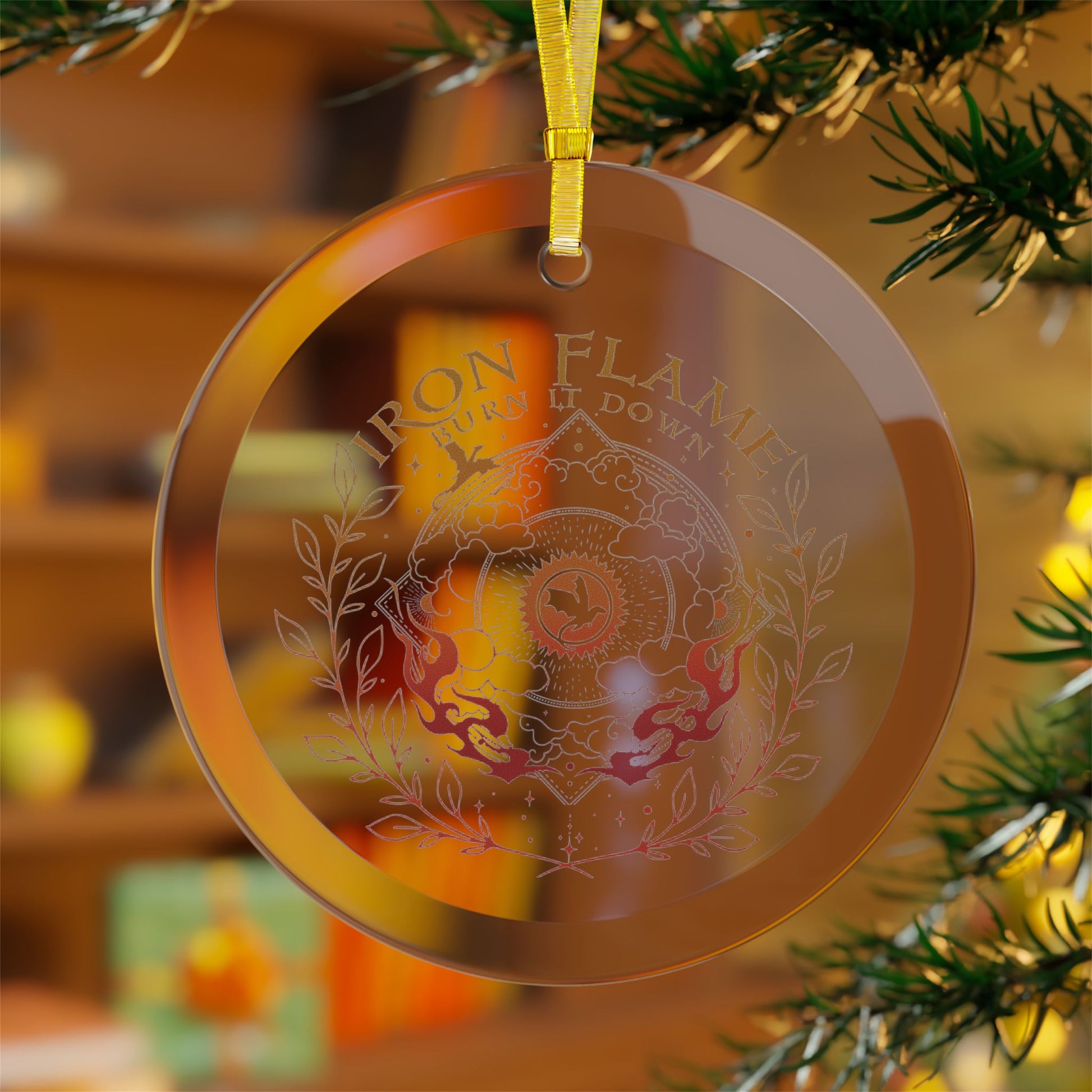 A round transparent acrylic Christmas ornament hanging from a yellow ribbon, featuring an etched design with the words 'IRON FLAMES BURN IT DOWN' and a central crest. The ornament is shown hanging in front of a blurred Christmas tree branch and warm holiday background.