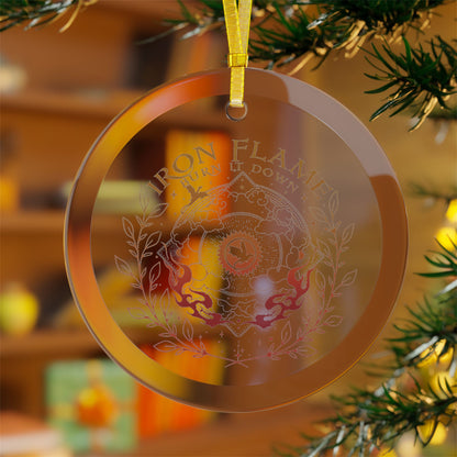 A round transparent acrylic Christmas ornament hanging from a yellow ribbon, featuring an etched design with the words 'IRON FLAMES BURN IT DOWN' and a central crest. The ornament is shown hanging in front of a blurred Christmas tree branch and warm holiday background.