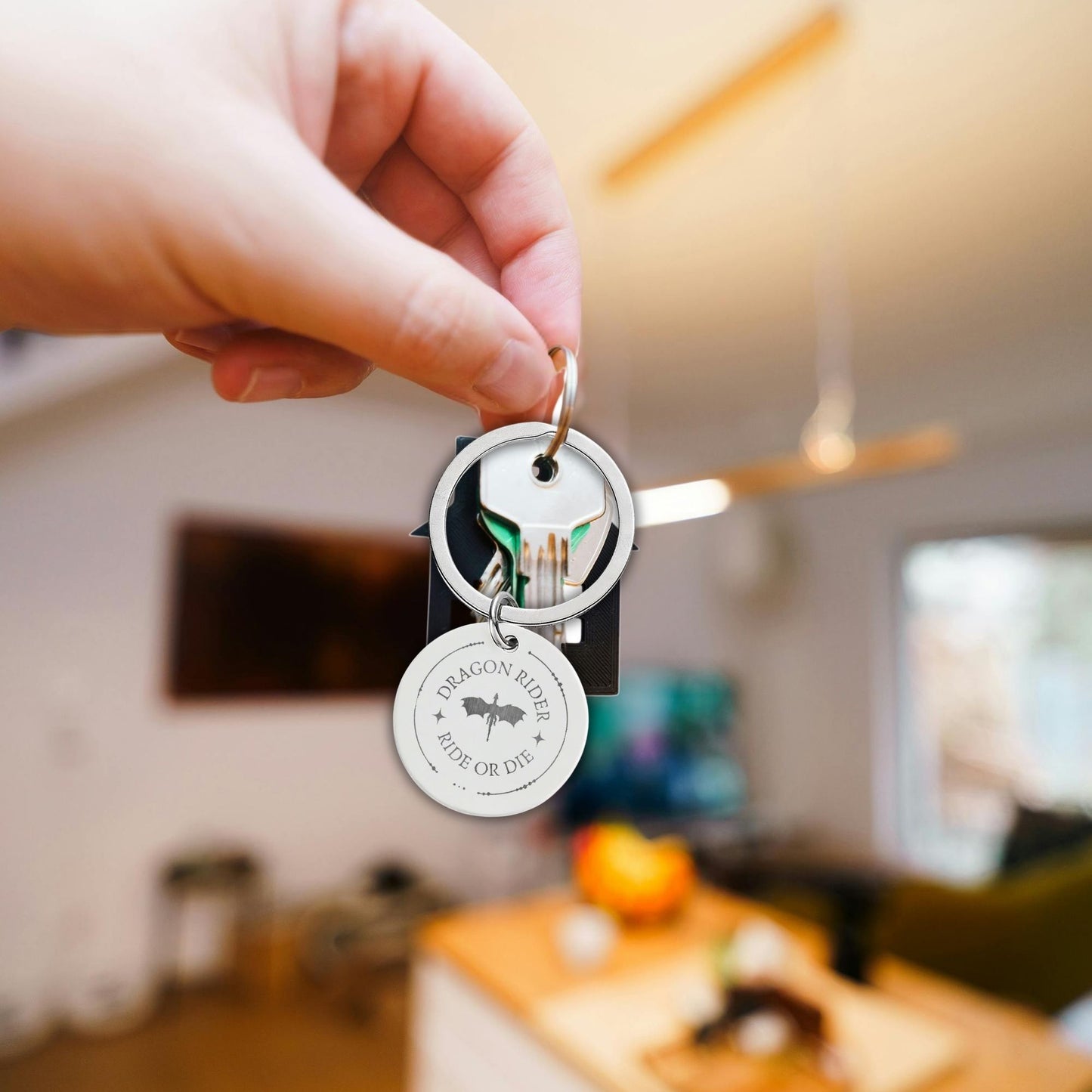 A hand holds a set of keys in a real indoor environment. The keys include a round silver metal keychain with a circular tag that reads 'DRAGON HIDE HIDE OR DIE' with a dragon graphic, and another round key tag.