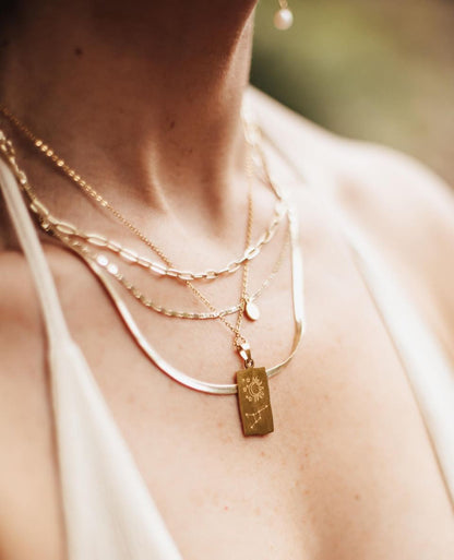 Close-up of a person wearing multiple gold necklaces with a focus on one with a tag featuring a zodiac sign.