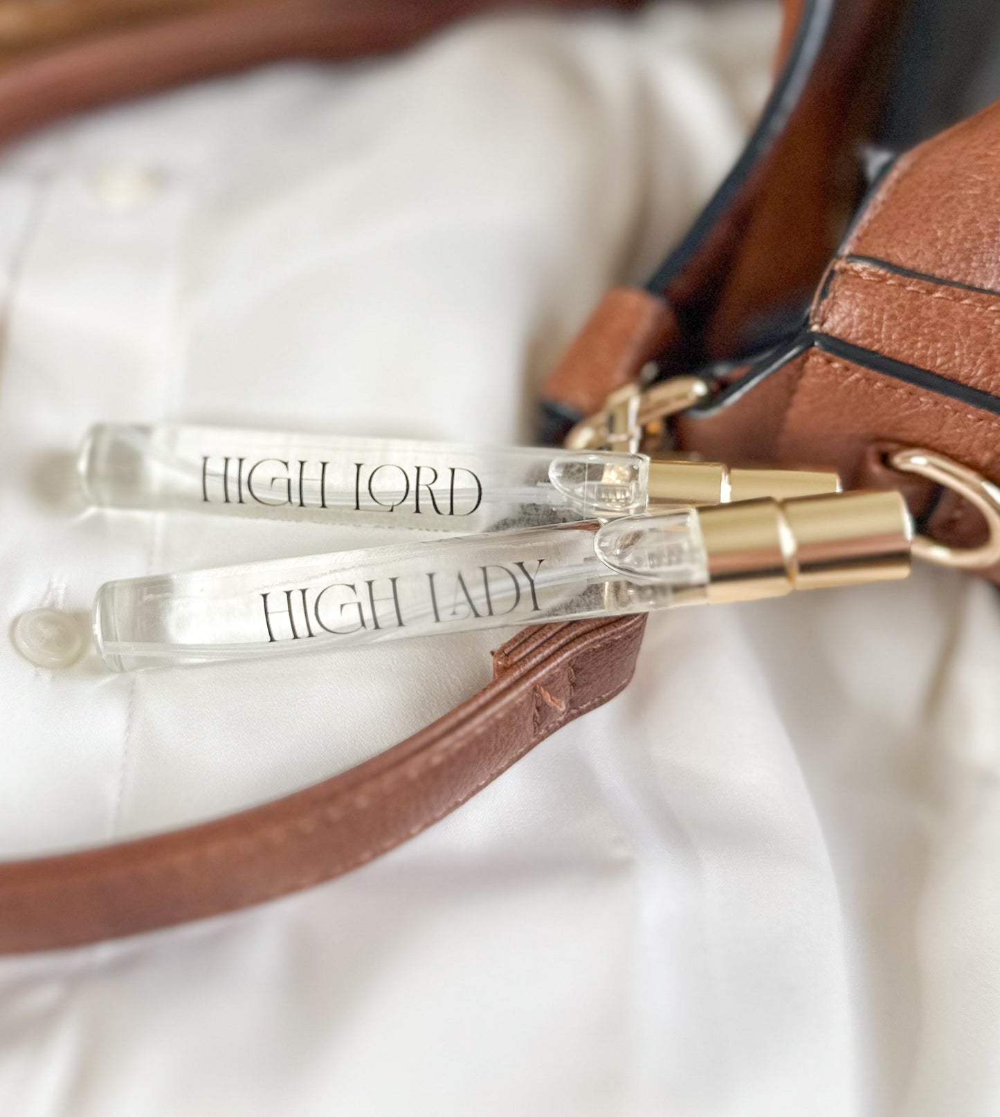 Two clear lip glosses with gold accents labeled 'HIGH LORD' and 'HIGH LADY' on a brown leather bag.