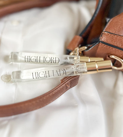 Two clear lip glosses with gold accents labeled 'HIGH LORD' and 'HIGH LADY' on a brown leather bag.