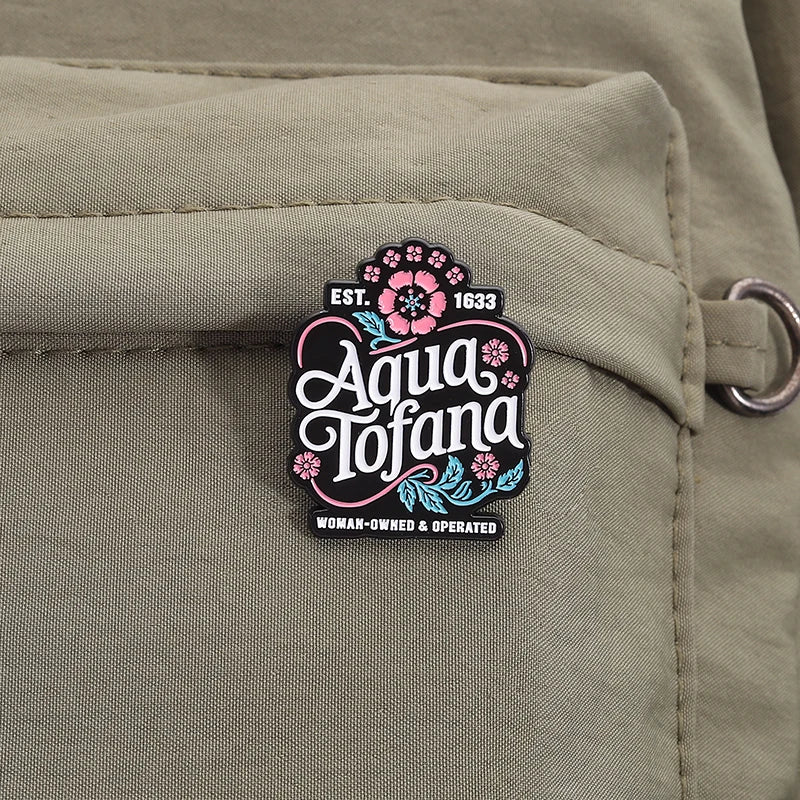 A close-up of a rectangular enamel pin with the brand name 'Aqua Tofana' and 'Est. 1633' on a khaki canvas background.