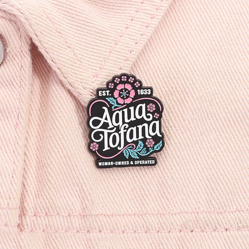 A close-up of a pink woven fabric, likely a denim jacket, featuring a decorative enamel pin that reads 'Aqua Tofana' with pink and teal floral details.