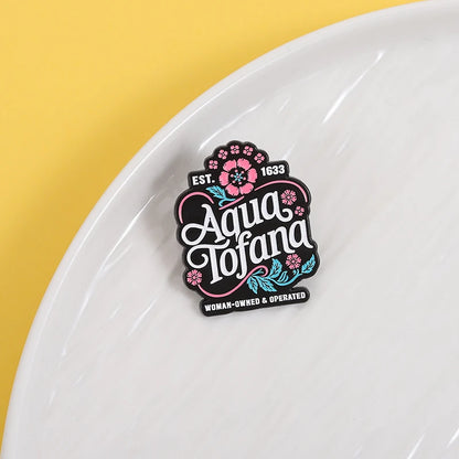 A small decorative enamel pin reading 'Aqua Tofana' with floral details and 'EST. 1633' and 'WOMAN-OWNED & OPERATED' on a white plate against a yellow background.