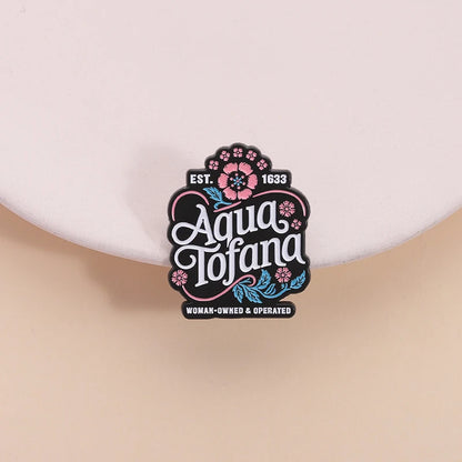 A small decorative enamel pin or badge reading 'Aqua Tofana' with floral accents and 'EST. 1833' and 'WOMAN-OWNED & OPERATED' on a pale pink background.