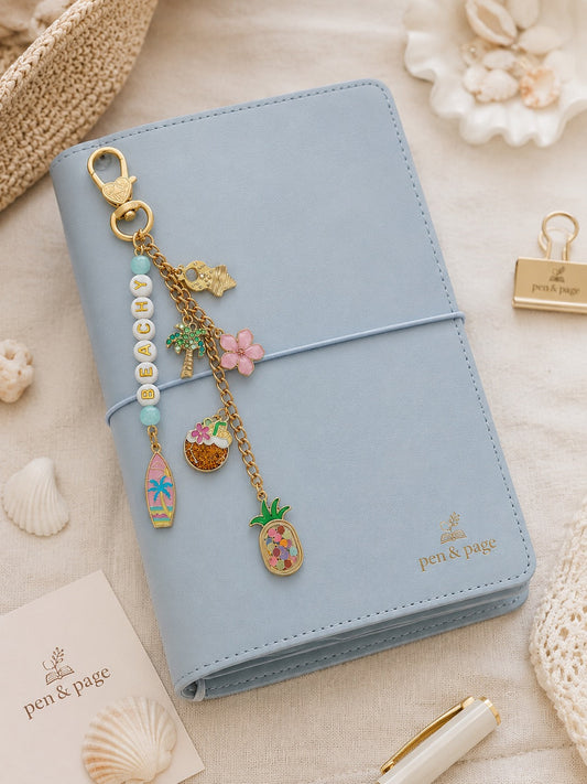 A light blue planner or notebook with a gold clasp bag charm featuring letter beads spelling "BEACHY", and beach inspired charms styled on a beige fabric background with seashells, a straw hat, and a pen.