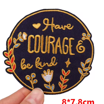 A circular embroidered patch held by a hand against a white background, featuring the text 'Have Courage & Be Kind' in gold thread with floral and leaf motifs in red, blue, and white. Red text at the bottom right reads '8*7.