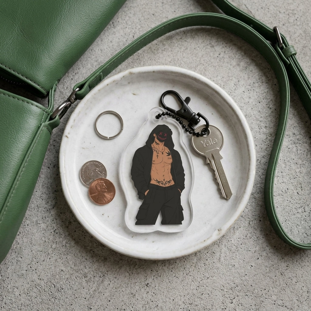A clear acrylic keychain featuring a cartoon-style illustrated figure in black clothing with a tan torso, attached to a black keyring and key. The keychain is displayed on a small white plate alongside coins, a ring, and a green leather strap on a gray textured surface.