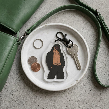 A clear acrylic keychain featuring a cartoon-style illustrated figure in black clothing with a tan torso, attached to a black keyring and key. The keychain is displayed on a small white plate alongside coins, a ring, and a green leather strap on a gray textured surface.