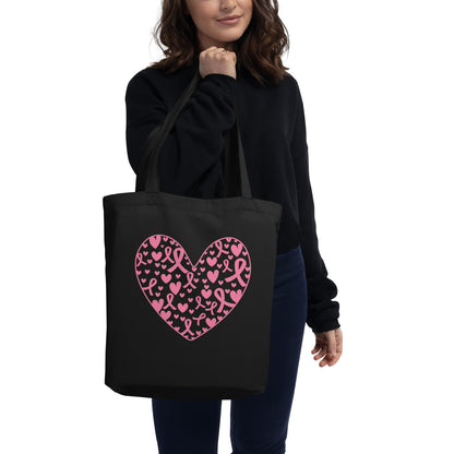 A woman wearing a black sweatshirt and blue jeans carries a black tote bag with a pink heart pattern made of breast cancer awareness ribbon and heart motifs, against a plain white background.