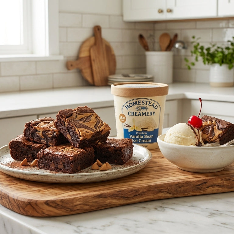 A tub of Homestead Creamery Vanilla Bean Brownie Ice Cream sits on a marble kitchen counter next to a plate of chocolate brownies and a bowl of vanilla ice cream with a cherry, set on a wooden serving board in a bright, modern kitchen.