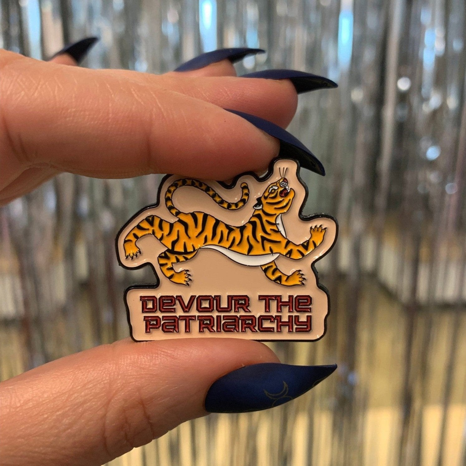 Pin with tiger design and 'Devour the Patriarchy' text held by a hand against a metallic background