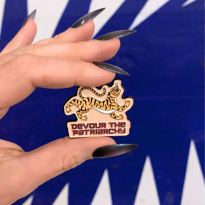Hand holding a pin with a tiger design and 'Devour the Patriarchy' text against a blue and white striped background.
