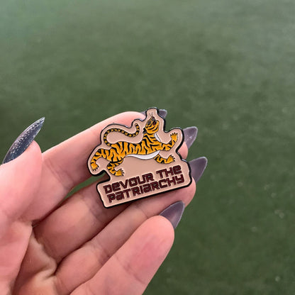 Hand holding a badge with a tiger design and 'Devour the Patriarchy' text against a green background