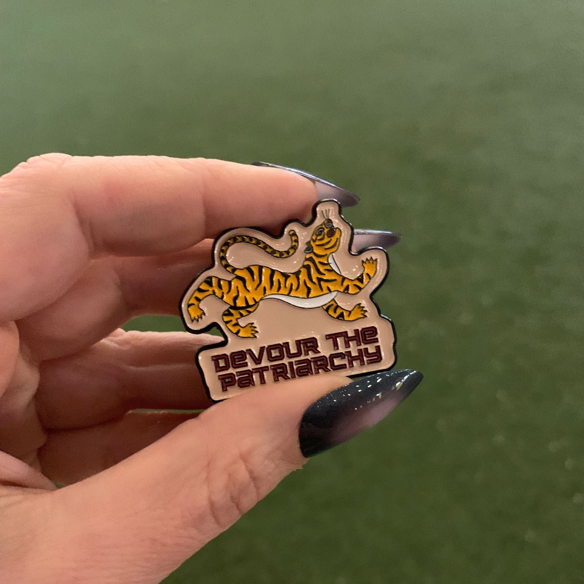 Hand holding a badge with a tiger design and 'Devour the Patriarchy' text against a green background