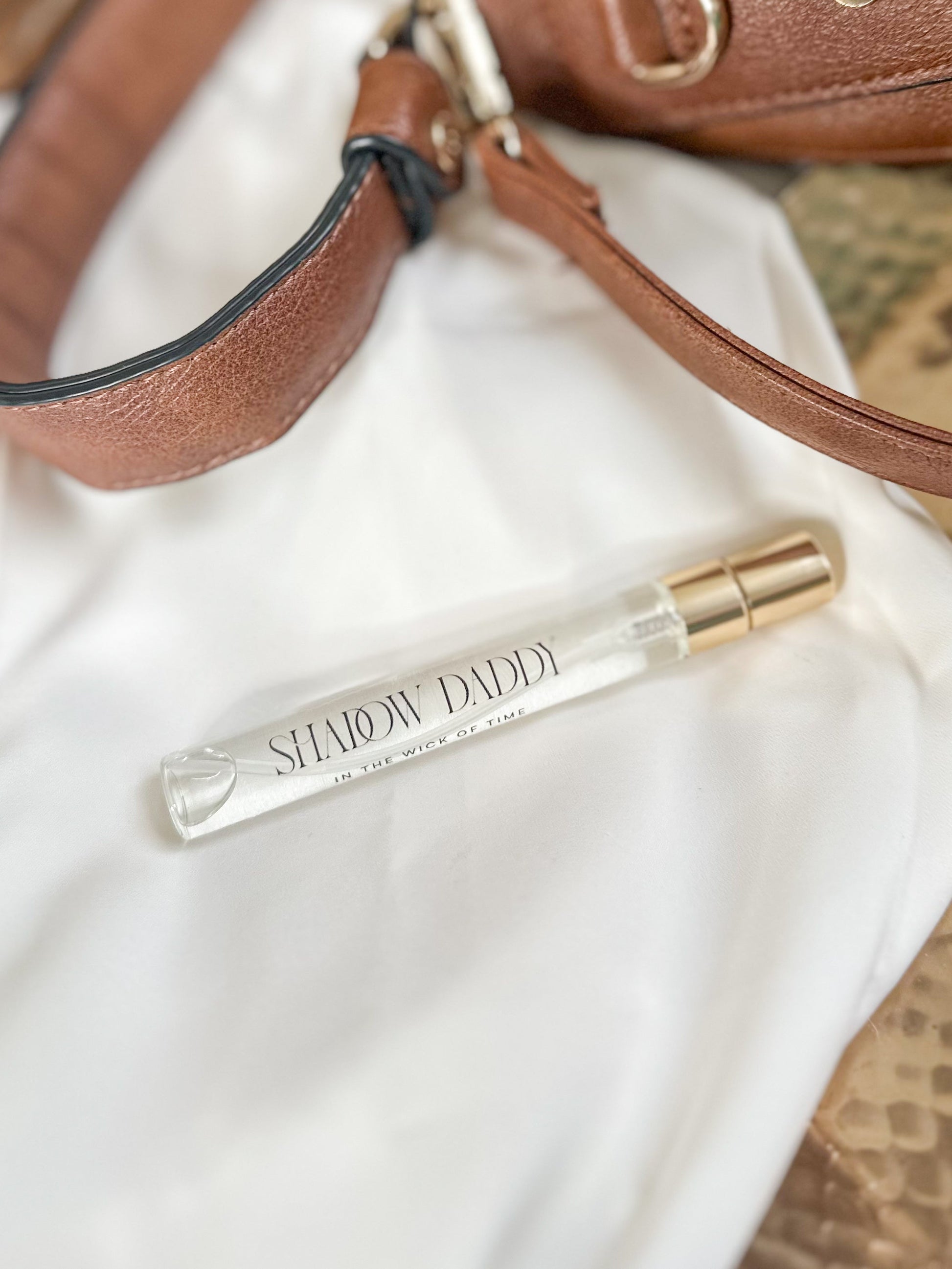 Shimmer Daddi perfume bottle on a white surface with a brown leather handbag in the background.
