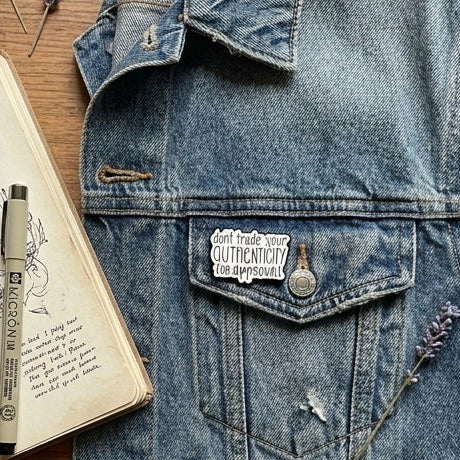 A styled flat-lay featuring a light blue denim jacket as the central product featuring a white enamel pin with black text that reads 'don't trade your authenticity for approval' surrounded by a notebook, pen, washi tape, glasses, a mug of coffee, and dried flowers on a wooden surface.