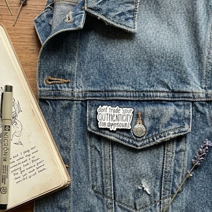 A styled flat-lay featuring a light blue denim jacket as the central product featuring a white enamel pin with black text that reads 'don't trade your authenticity for approval' surrounded by a notebook, pen, washi tape, glasses, a mug of coffee, and dried flowers on a wooden surface.