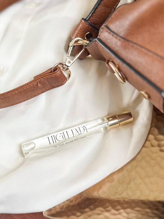 Clear Highclad perfume bottle with gold cap on a white surface with brown leather bag.