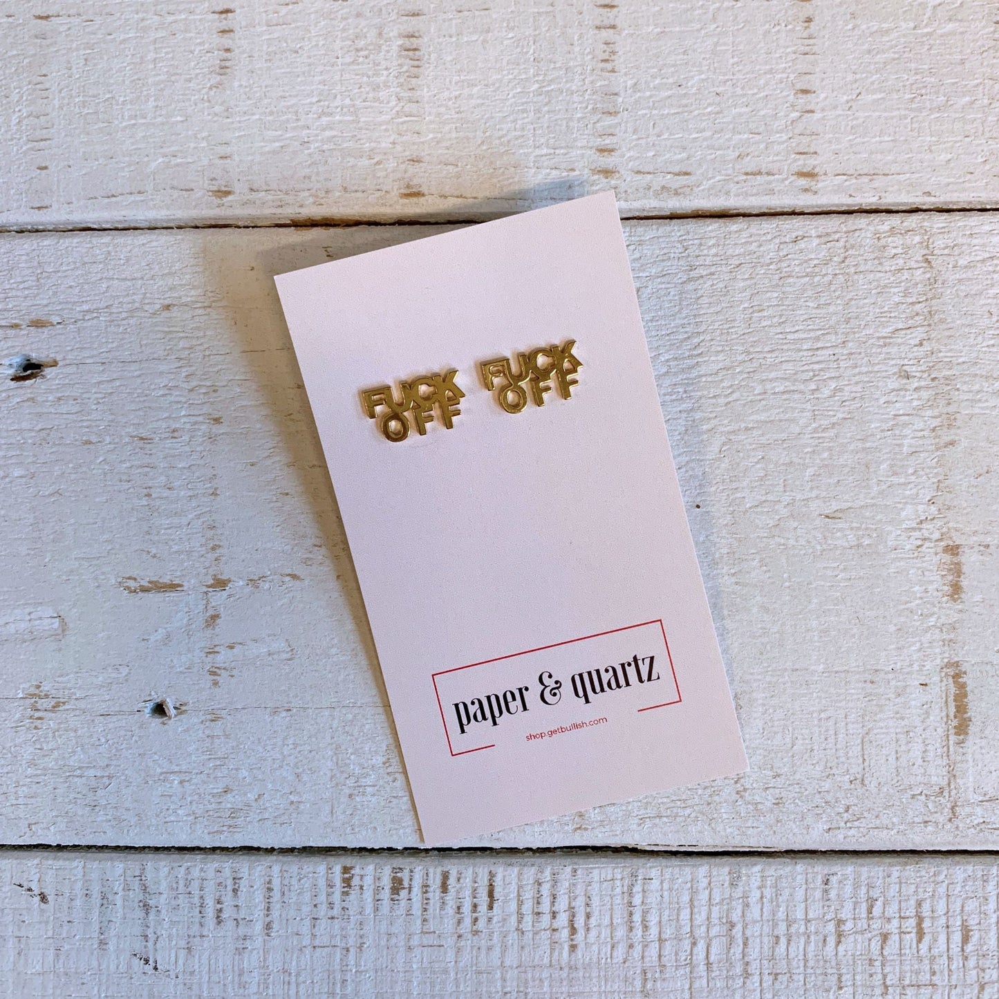 Gold 'F*** Off' pins on a pink card with 'paper & quartz' branding on a wooden surface.