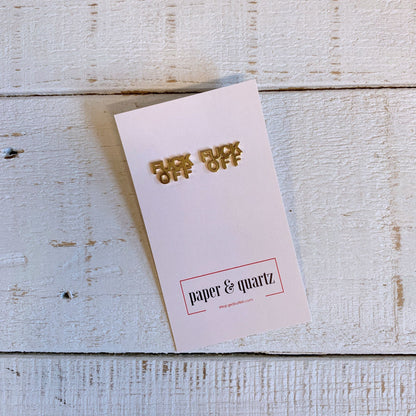 Gold 'F*** Off' pins on a pink card with 'paper & quartz' branding on a wooden surface.