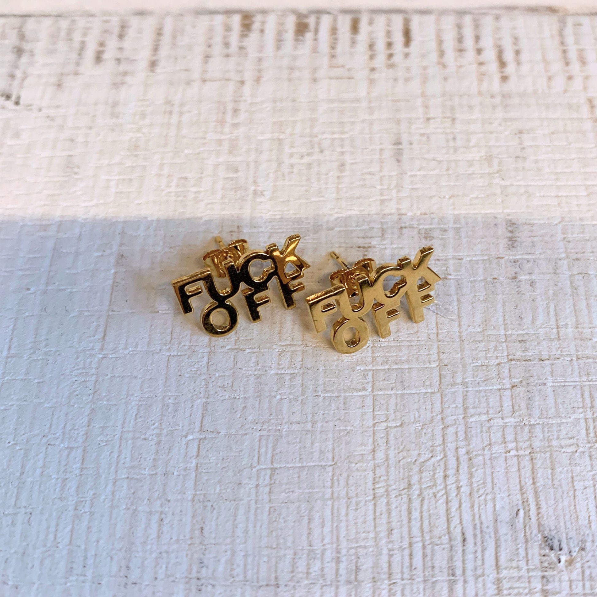 Gold earrings with 'Foxy' design on a textured beige background