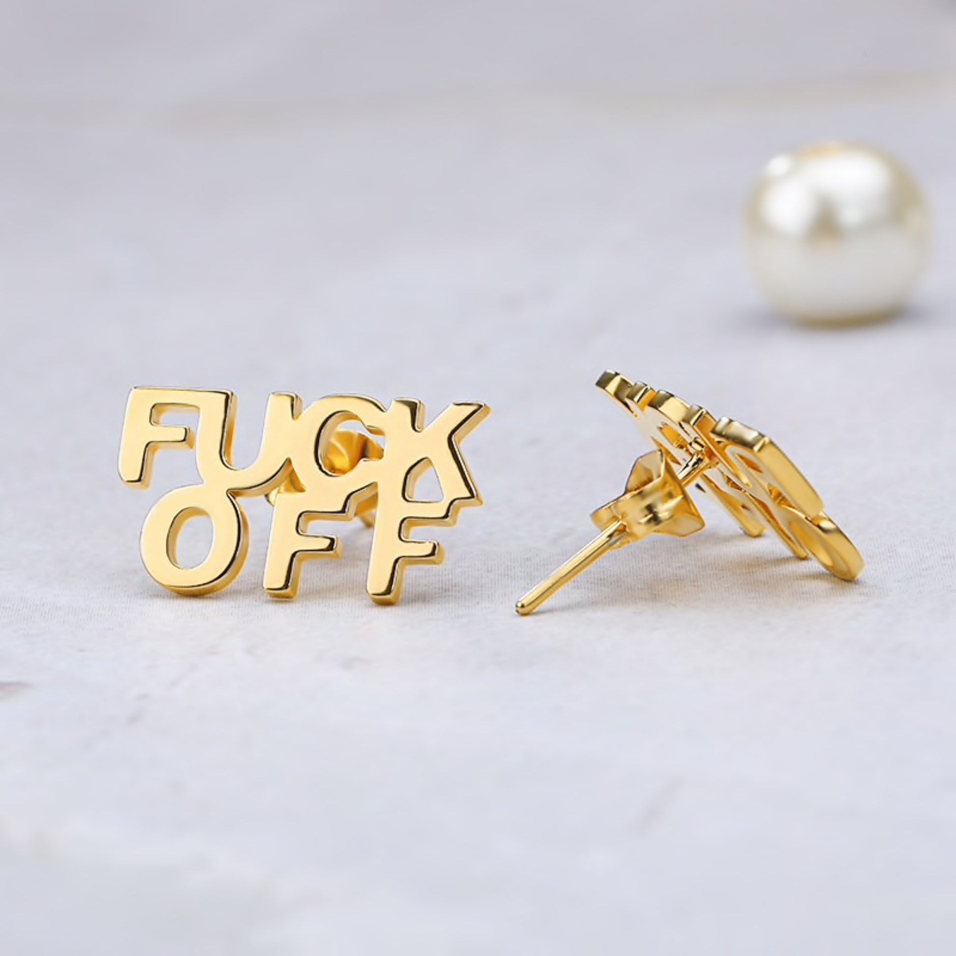 Gold 'FUCK OFF' earrings on a light gray background