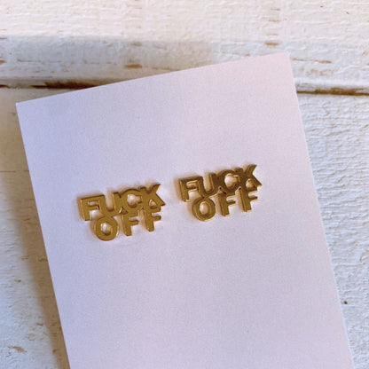 Gold 'FUCK OFF' earrings on a white card with a wooden surface background