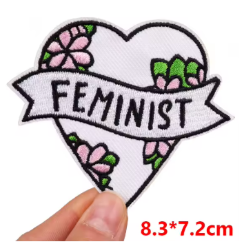 A heart-shaped embroidered patch with the word 'FEMINIST' on a banner, decorated with pink flowers and green leaves, held by a hand against a white background with red size text reading 8.3*7.