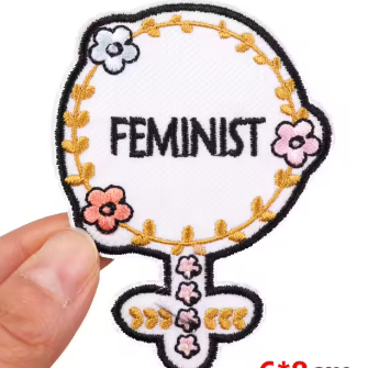 A hand holds a white embroidered patch shaped like a feminist symbol, with black text reading 'FEMINIST,' a gold floral border, and pink and white flowers. A red size label in the corner reads 6*8cm.