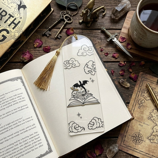 A clear acrylic bookmark with a gold tassel and printed with a dragon illustration, and the phrase 'FLY OR DIE' resting on an open book in a styled tabletop scene with keys, a cup of tea, dried flowers, and a map.
