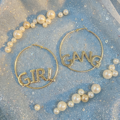 Gold hoop earrings with 'GIRL' and 'GANG' text on a glittery blue background with pearls.
