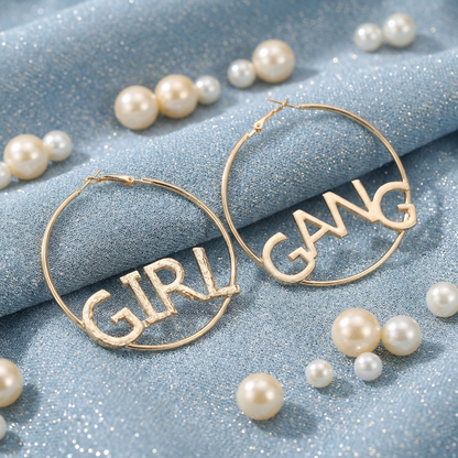 Gold hoop earrings with 'GIRL GANG' text on a blue fabric background with pearls.