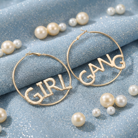 Gold hoop earrings with 'GIRL GANG' text on a blue fabric background with pearls.