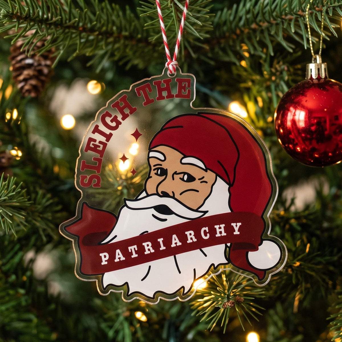 A custom Christmas ornament featuring a cartoon Santa with the text 'SLEIGH THE PATRIARCHY' hanging on a decorated Christmas tree with lights and ornaments.