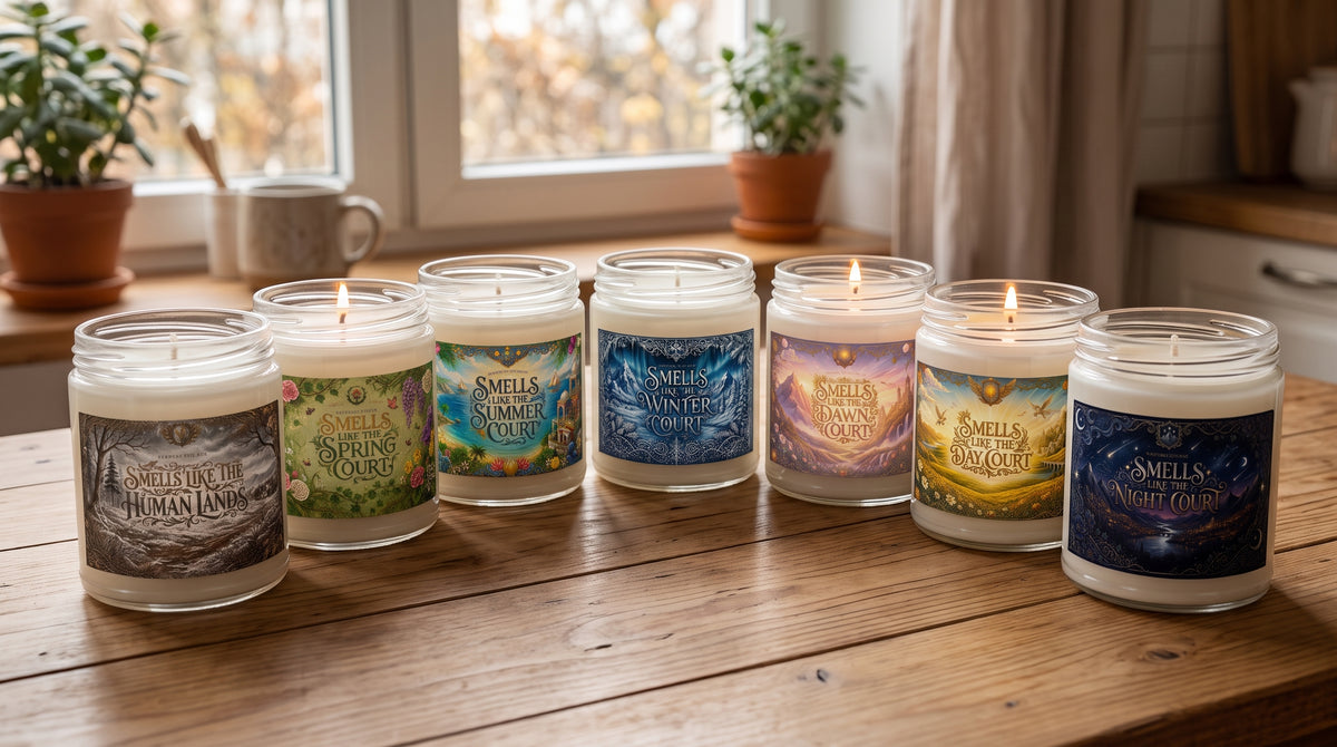 Six candles in clear glass jars with colorful illustrated labels are arranged on a wooden table in a warm, sunlit room near a window.