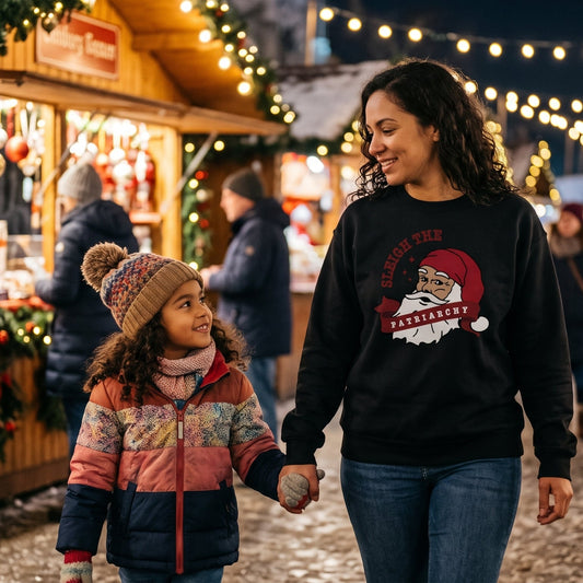 A woman and a child holding hands walking through a festive Christmas market at night, with wooden stalls decorated with lights and greenery in the background.